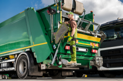 A man hanging off the back of a trash pick up truck