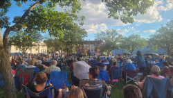 A large audience sitting in chairs in a park listening to live music. 