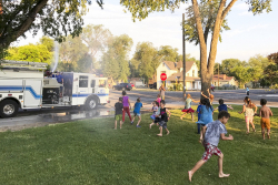 Fire truck in the background and kids running through water in a park.